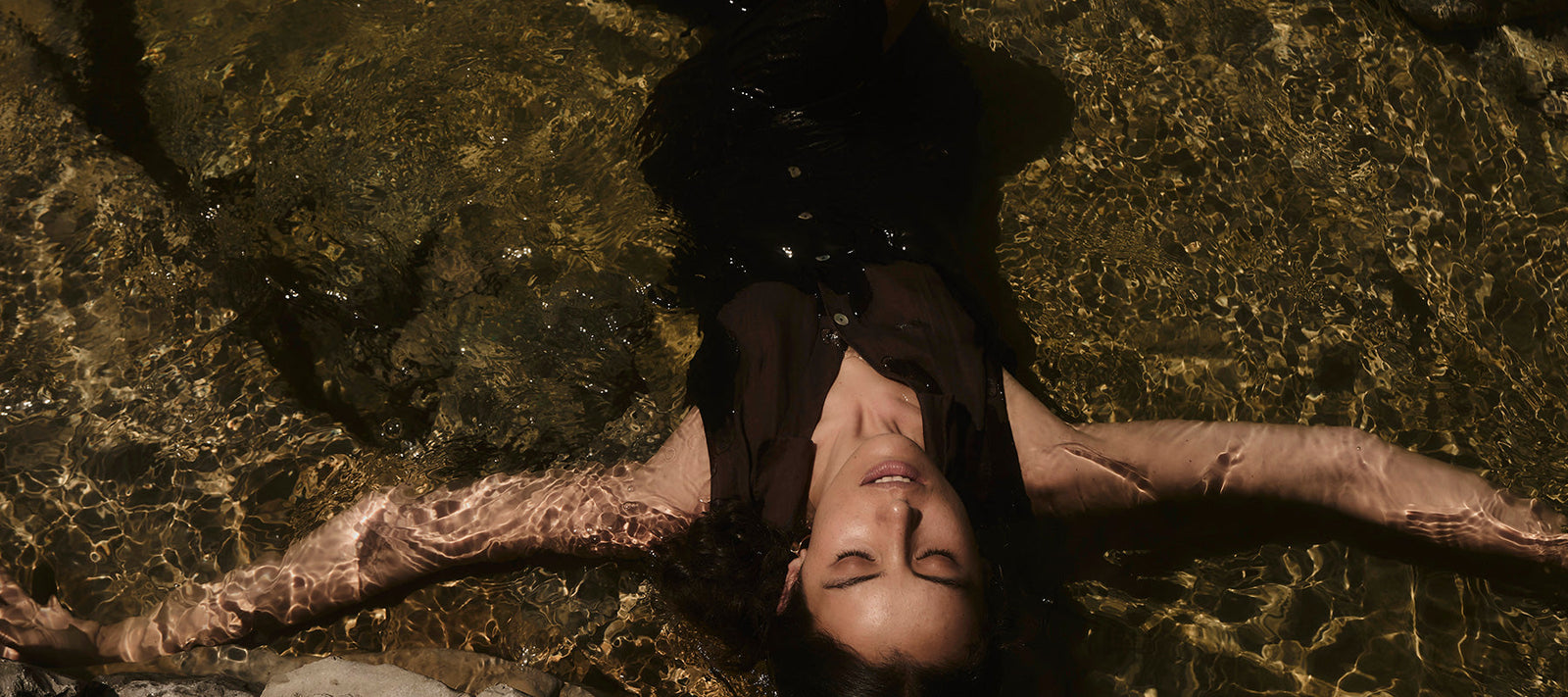 Woman with dark brown dress lies in water with closed eyes